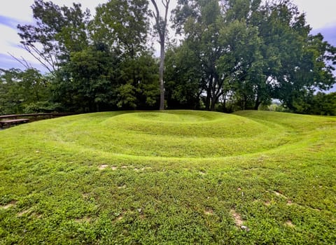 Side view closeup of the coiled tail of the snake at Serpent Mound in Ohio, the largest effigy mound in the world. Ridges of the prehistoric Native American engineering earthwork art are clearly visible covered in green grass with trees and dramatic sky background and no people.