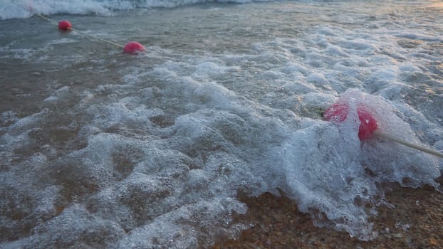 Buoys on a rope near sea water. The buoys are pink restraints to alert people to the depth of the water. Rescue of the drowning. Delimiting a place on a sandy beach between hotels. Wave with bubbles