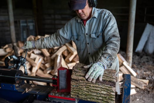Caucasian senior man splits wood with a hydraulic log splitter. He wears a denim work shirt, work gloves and ear protection. Defocused wood pile in background