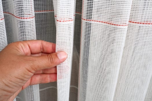 White coarse cotton or linen curtains made of weaves of thin threads. Simple compact economical apartment interior. Window decoration in the room. A woman's hand strokes the tulle, shows the quality