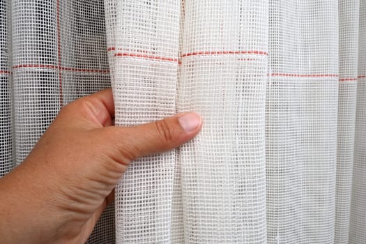 White coarse cotton or linen curtains made of weaves of thin threads. Simple compact economical apartment interior. Window decoration in the room. A woman's hand strokes the tulle, shows the quality