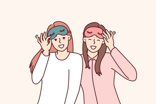 Friendly girls in pajamas with sleep masks are preparing to relax in comfortable bedroom with soft bed. Happy women use eye masks to protect themselves from sunlight during daytime naps or siestas