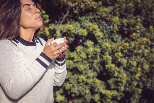 A Latina woman sits on a wooden bench in the garden, enjoying the fresh air while taking some time for herself. She holds a cup of coffee and smiles with a relaxed expression, enjoying a moment of tranquility.