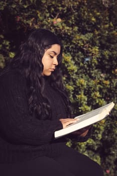 young woman, seated attentively reading a book in the garden vertical half-body side view photo