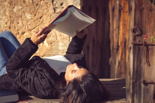 close-up from behind of latin woman reading a big book in the sun on a wooden table in the garden of the house