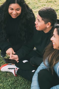 three individuals of Hispanic-Latino ethnicity sitting on the lush green grass of a park, with smart device. Relaxed and smiling expressions on their faces they are enjoying a moment of leisure amidst the hustle and bustle of everyday life.