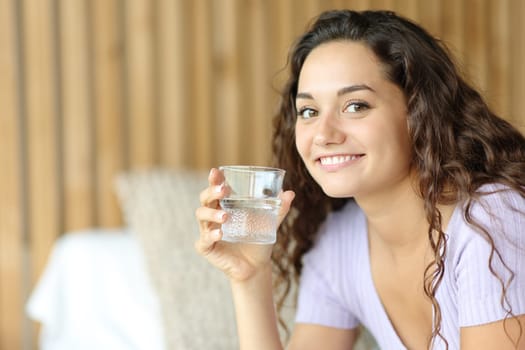 Happy woman holding water glass looks at you sitting on a bed