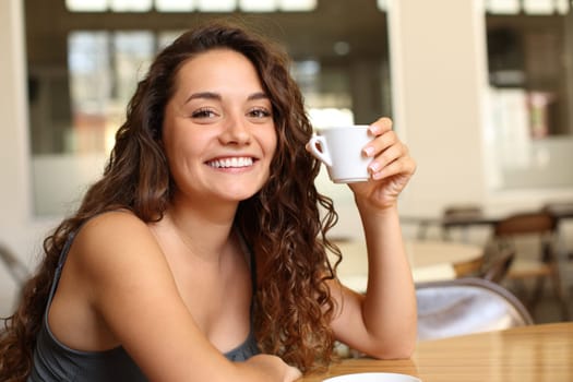 Happy woman holding coffee mug looking at you sitting in a bar and smiling