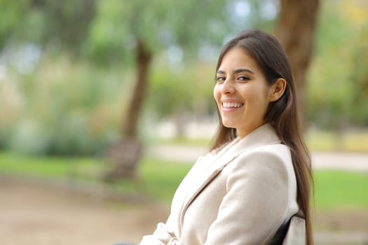 Happy woman in winter looking at camera sitting on a bench in a park