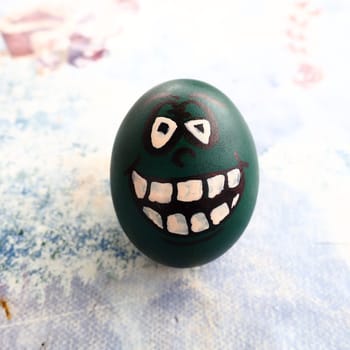 Easter egg dyed green with a painted laughing face. Cool grimace with eyes and big white teeth. Scary mug for Halloween. Emoticon for Easter. Light abstract background
