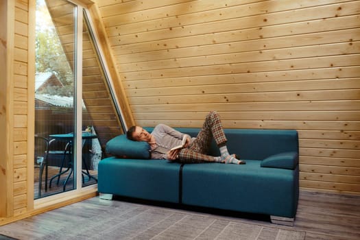 Middle aged man has retired to a country wooden cabin and is reading a book lying on the sofa