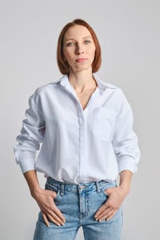 Positive middle aged woman in white shirt and blue jeans put thumbs in pockets over grey background