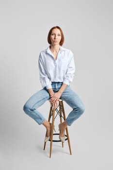 Lifestyle portrait of middle aged caucasian redhead woman in white shirt and blue jeans sitting on tall stool
