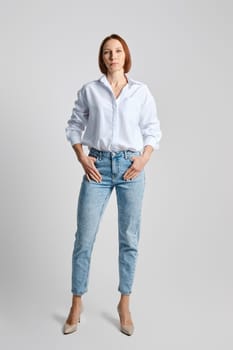 Relaxed middle aged woman wearing white shirt and blue jeans over grey background