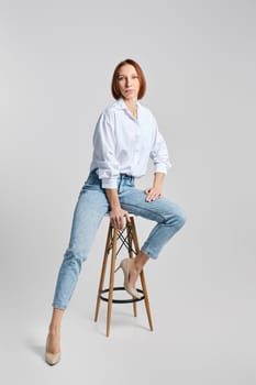 Casual portrait of middle aged redhead woman in white shirt and blue jeans sitting on tall stool and stretching one leg to the side