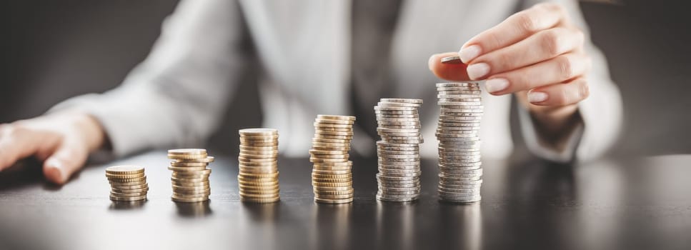 Woman sitting at the table and putting coin on the largest stack of the money. Concept of the wages and financial income in business.