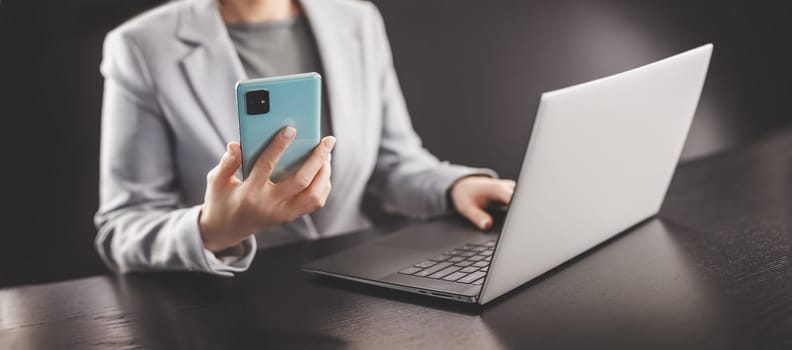Woman in the office holding in her hands mobile phone and typing on the laptop. Concept of the e-commerce, business and surfering in network.