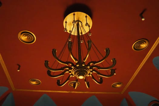 Theatrical golden chandelier under the ceiling of the theater hall. Solemn and rich interior lighting option. Theatrical atmosphere. Interior in red, burgundy and gold. Energy saving and safety