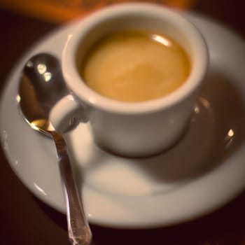 Cup of coffee in cafe. Anamorphic bokeh effect. Tilt-shift photo. Hot coffee in a white cup with a saucer and a spoon. The coffee is getting cold. Aroma of coffee beans. Label or advertisement. Foam