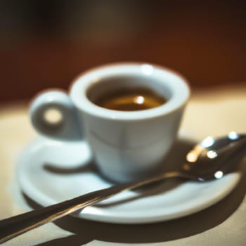 Cup of coffee in cafe. Anamorphic bokeh effect. Tilt-shift photo. Hot coffee in a white cup with a saucer and a spoon. The coffee is getting cold. Aroma of coffee beans. Label or advertisement. Foam