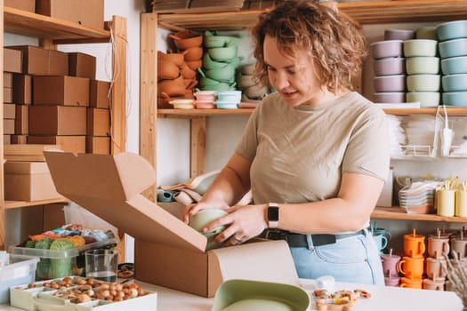 Smiling concentrated curly woman packing and wrapping shipment with silicone baby dish in cardboard box for delivery. Wooden rattle toy, trinket decoration nearby on desk. Workshop store, work at home