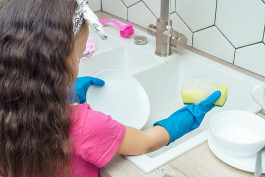Little girl in protective gloves washing up white plate with foamy dishwashing sponge and dish soap in kitchen sink. Portrait of child tidying up kitchen back view. Home cleaning concept.