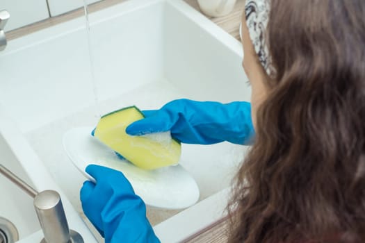 Little girl in protective gloves washing up white plate with foamy dishwashing sponge and dish soap in kitchen sink. Portrait closeup of child tidying kitchen from above view. Home cleaning concept.