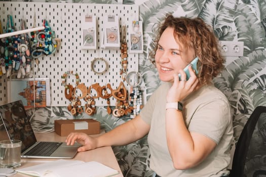 Smiling concentrated business woman talking smartphone, working on laptop in workshop. Baby healthcare accessories, silicone eco rattles, teethers, pacifiers, hanging on white wall. Home office
