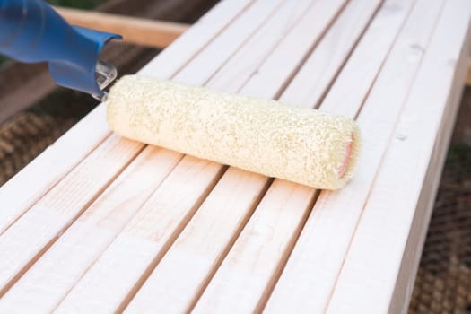 Side view of white soft paint roller applicator for coloring walls and floor, brushing wooden bench outdoors. Construction site work, renewal and renovation. White color palette, house repairment
