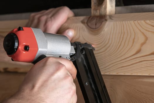 Close up cropped carpenter male hands using pneumatic nailer, stapler gun for wooden timber plank. Making layout on wooden plank. Builder handwork and wood repair technology. Craftsmanship, handcraft