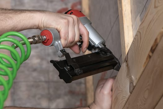 Unrecognizable cropped male hands using pneumatic nailer, nail gun with spiral cable for wooden timber board. Make layout on wooden plank. Clipping and scoring metal nails and staples. Low angle view
