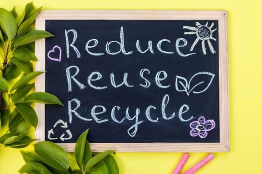 Chalk board Reduse Reuse Recycle sign on a yellow background with green leaves and chalk pieces. Top view