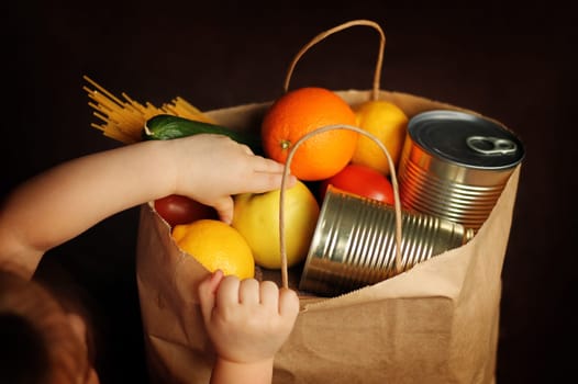Little girl puts products in a paper bag on a brown background. A child takes food out of a food delivery service package. Canned food, pasta, vegetables, fruits. Contactless delivery.
