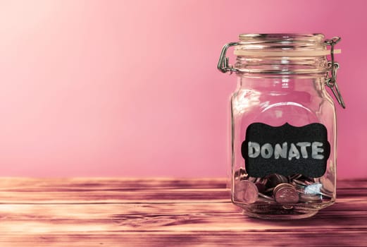 Glass jar with coins with chalk tag Donate on a pink background, donation and charity concept. Copy space.