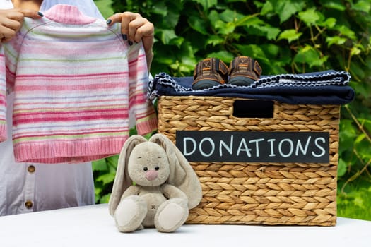 Hands putting a kids sweater in a box for clothing donations on a greenery background. Charity and donation concept, copy space.