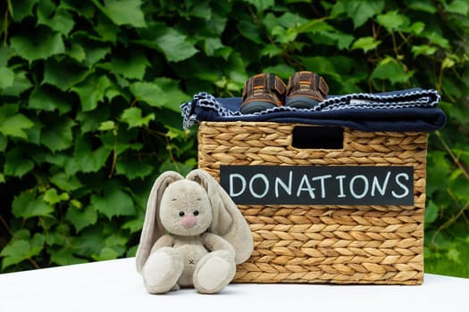 Donation box for clothing donations with children shoes and toy hare on a greenery background. Charity and donation concept. Copy space.