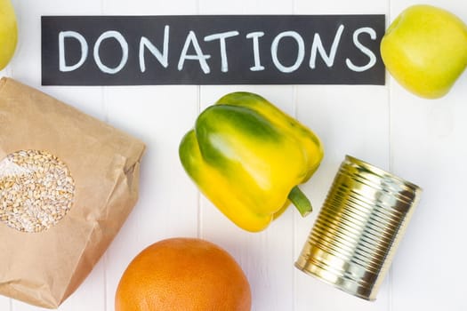 Cereals, canned food, milk, vegetables and fruits with a chalk Donations sign on a white wooden table. Flat lay. Charity and donation concept.