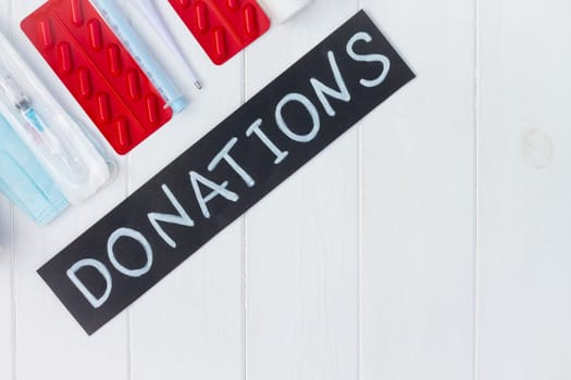 Medications, masks, gloves, syringes, thermometer with a chalk Donations sign on a white wooden table. Flat lay. Copy space. Charity and donation concept.