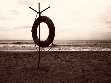 Lifebuoy on a sandy beach. Orange circle on a pole to rescue people drowning in the sea. Rescue point on the shore. Sky and sea in the background. Sepia monochrome.