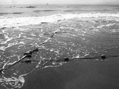 Buoys on a rope near sea water. The lifebuoys restraints to alert people to the depth of the water. Delimiting a place on a sandy beach between hotels. Wave with bubbles. Black white photo monochrome.