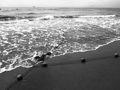 Buoys on a rope near sea water. The lifebuoys restraints to alert people to the depth of the water. Delimiting a place on a sandy beach between hotels. Wave with bubbles. Black white photo monochrome.