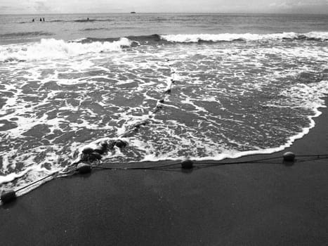 Buoys on a rope near sea water. The lifebuoys restraints to alert people to the depth of the water. Delimiting a place on a sandy beach between hotels. Wave with bubbles. Black white photo monochrome.