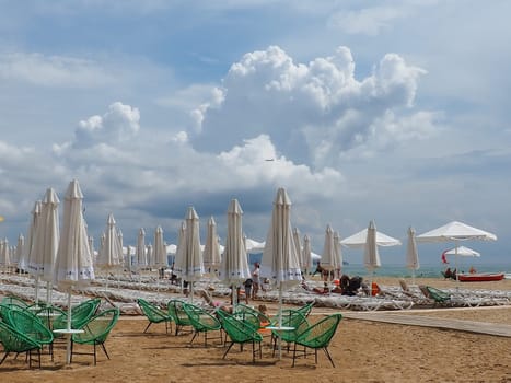 Anapa, Russia, August 15, 2021 Sun loungers and umbrellas on the sandy sea beach after the rain. Beach season. Beach cafe with wicker chairs. Tourists walking people. A storm warning. Cumulus clouds.