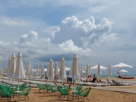 Anapa, Russia, August 15, 2021 Sun loungers and umbrellas on the sandy sea beach after the rain. Beach season. Beach cafe with wicker chairs. Tourists walking people. A storm warning. Cumulus clouds.
