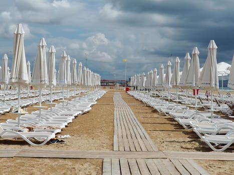Wet sunbeds and umbrellas on the sea beach during heavy rain storm. Rolled up parasol on the beach against dark cloudy sky. Invasion of a typhoon, cyclone or storm on the sea or ocean. Beach season