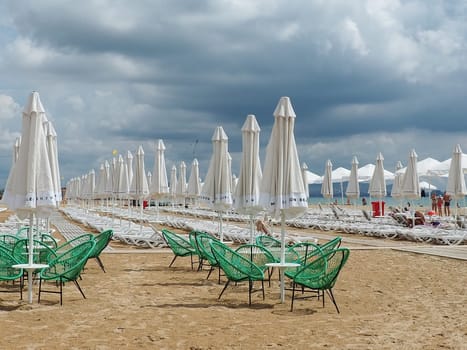 Anapa, Russia, August 15, 2021 Sun loungers and umbrellas on the sandy sea beach after the rain. Beach season. Beach cafe with wicker chairs. Tourists walking people. A storm warning. Cumulus clouds.