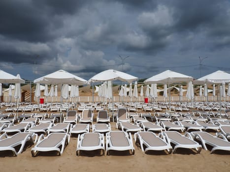 Wet sunbeds and umbrellas on the sea beach during heavy rain storm. Rolled up parasol on the beach against dark cloudy sky. Invasion of a typhoon, cyclone or storm on the sea or ocean. Beach season