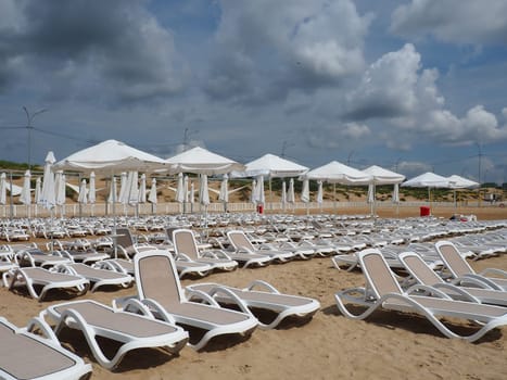 Anapa, Russia, August 15, 2021 Wet sunbeds and umbrellas on the sea beach during heavy rain storm. Rolled up parasol on the beach against dark cloudy sky. Invasion of a typhoon, cyclone or storm.
