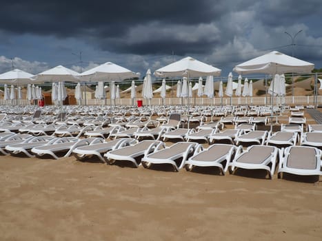 Wet sunbeds and umbrellas on the sea beach during heavy rain storm. Rolled up parasol on the beach against dark cloudy sky. Invasion of a typhoon, cyclone or storm on the sea or ocean. Beach season