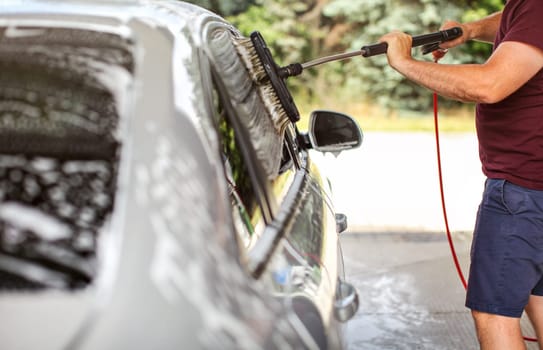Young man in shorts and t shirt washing his car in self serve carwash, cleaning side windows with brush.